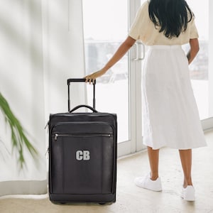 Könnte beinhalten: Schwarzer Rollkoffer aus Leder mit einem silbernen Reißverschluss und einem silbernen Monogramm "CB" auf der Vorderseite.