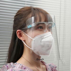 May include: A clear plastic face shield with a white face mask underneath. The face shield has a clear plastic frame and a clear plastic visor. The face shield has a small blue tab on the side. The face mask is white and has a white strap. The face shield has the word "Brenda" written on the bottom.