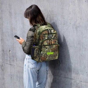 May include: A camouflage-patterned backpack with multiple compartments and a side pocket for a water bottle. The backpack has the word "CENA" printed in bright green. The backpack is worn by a person wearing a jacket and jeans.