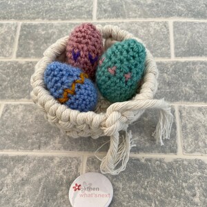 May include: Three crocheted Easter eggs in a white crocheted basket. The eggs are blue, pink, and green with embroidered details.