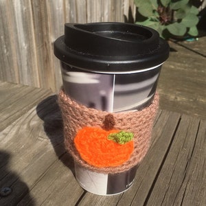 Puede incluir: Una taza de viaje negra con un dise&ntilde;o de manzana naranja tejida en el acogedor. La taza tiene una tapa negra y un dise&ntilde;o estampado blanco y gris.