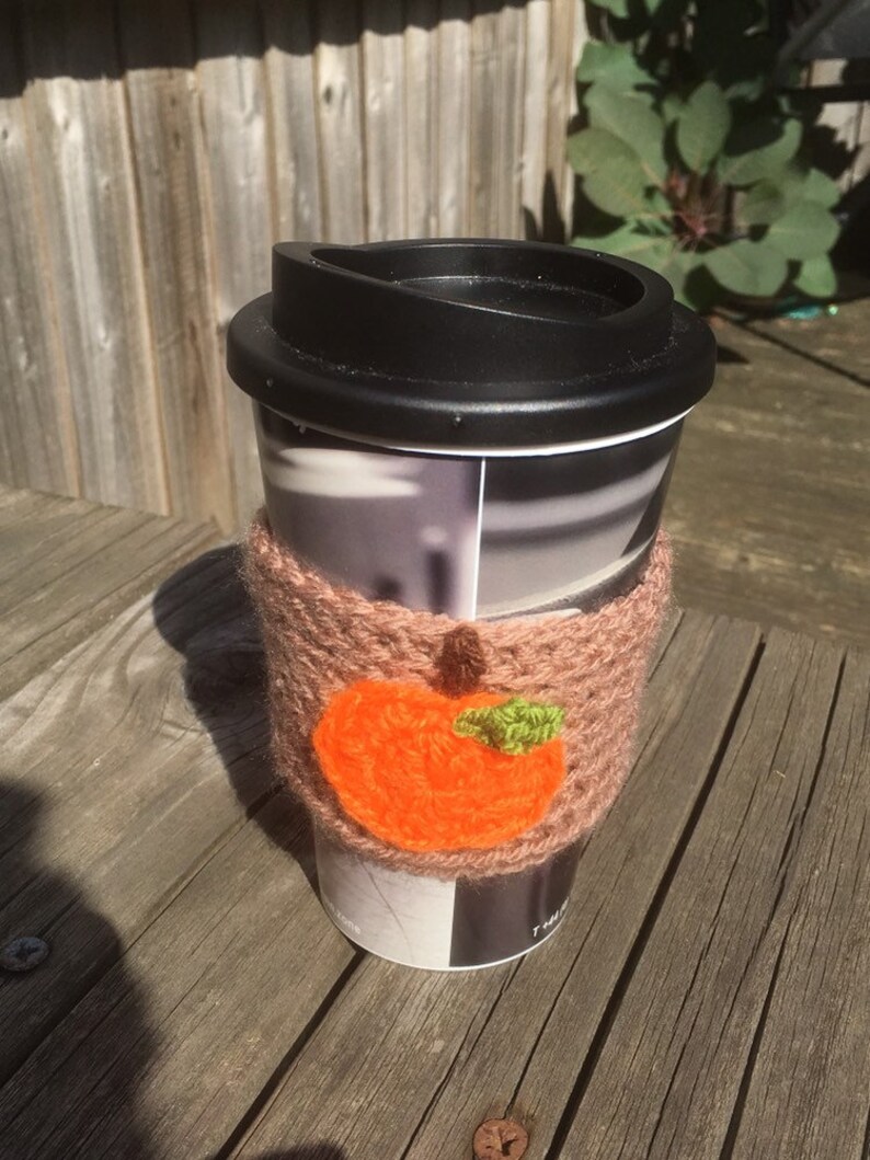Puede incluir: Una taza de viaje negra con un dise&ntilde;o de manzana naranja tejida en el acogedor. La taza tiene una tapa negra y un dise&ntilde;o estampado blanco y gris.