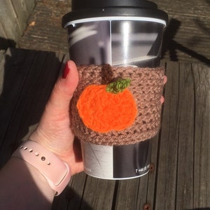 Puede incluir: Un calentador de taza de caf&eacute; de ganchillo marr&oacute;n con una aplicaci&oacute;n de calabaza naranja. El calentador est&aacute; en una taza de viaje con una tapa negra.