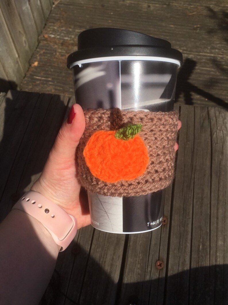 Puede incluir: Un calentador de taza de caf&eacute; de ganchillo marr&oacute;n con una aplicaci&oacute;n de calabaza naranja. El calentador est&aacute; en una taza de viaje con una tapa negra.