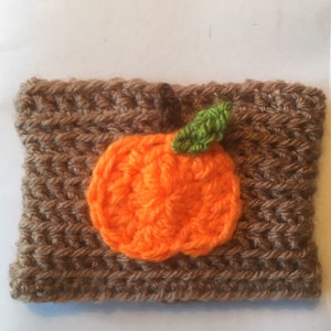 Puede incluir: Un calentador de taza de caf&eacute; de ganchillo marr&oacute;n con un dise&ntilde;o de calabaza de ganchillo naranja y una hoja de ganchillo verde.