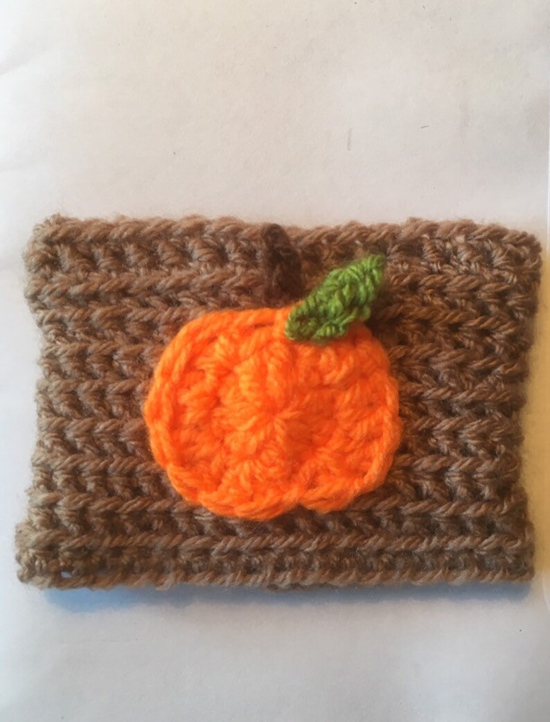 Puede incluir: Un calentador de taza de caf&eacute; de ganchillo marr&oacute;n con un dise&ntilde;o de calabaza de ganchillo naranja y una hoja de ganchillo verde.