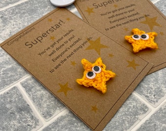 May include: Two brown cards with the text "Congratulations Superstar! You've got your results, well done to you! Everyone's excited, to see the next thing you do." Each card has a yellow crocheted star with googly eyes attached.