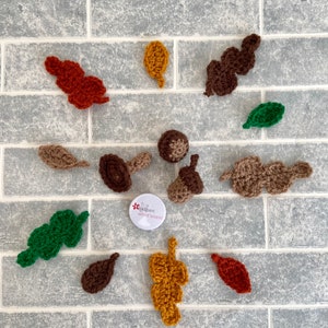 May include: A collection of crocheted leaves and acorns in various colours, including brown, green, orange, and yellow. The leaves are arranged on a grey brick background. A small button with the text "Crochet what's next" is also visible.