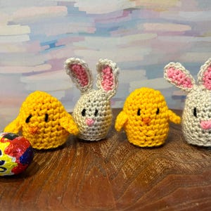 May include: Three crocheted Easter decorations: two yellow chicks and one white bunny. The bunny has pink ears and a pink nose. The chicks have black eyes and orange beaks. A chocolate egg is in the foreground.