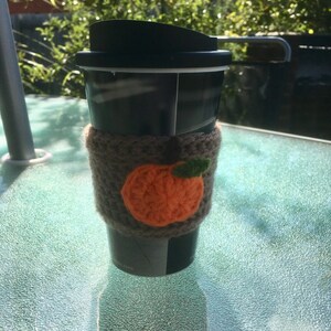 Puede incluir: Una taza de viaje negra con un acogedor de punto marr&oacute;n que presenta una calabaza naranja de ganchillo con un tallo verde.