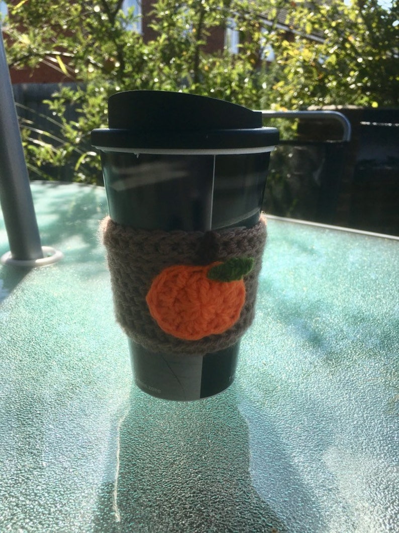 Puede incluir: Una taza de viaje negra con un acogedor de punto marr&oacute;n que presenta una calabaza naranja de ganchillo con un tallo verde.