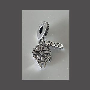May include: Silver charm featuring a detailed depiction of the Millennium Falcon spaceship from Star Wars. The charm has a small, dangling charm shaped like a lightsaber.