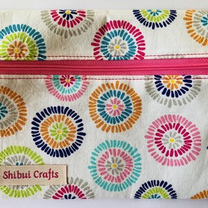 Puede incluir: Una bolsa blanca con cremallera rosa y un estampado floral colorido. La bolsa tiene una etiqueta que dice "Shibui Crafts".