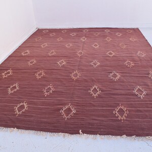 May include: A large, rectangular maroon rug with a repeating diamond pattern in a lighter beige color. The rug has a textured appearance with horizontal lines and a fringe along the bottom edge. The rug is suitable for a living room or bedroom.