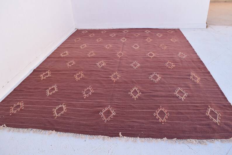 May include: A large, rectangular maroon rug with a repeating diamond pattern in a lighter beige color. The rug has a textured appearance with horizontal lines and a fringe along the bottom edge. The rug is suitable for a living room or bedroom.