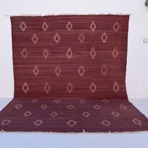 May include: A burgundy-colored rug with a diamond pattern in a lighter shade. The rug is rectangular with a fringed edge and is displayed against a white wall. The diamond pattern is evenly spaced across the rug's surface.