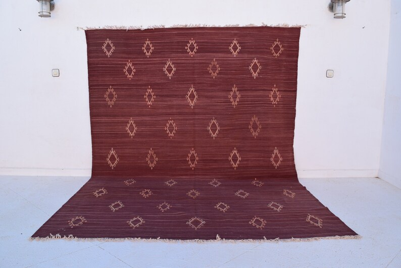 May include: A burgundy-colored rug with a diamond pattern in a lighter shade. The rug is rectangular with a fringed edge and is displayed against a white wall. The diamond pattern is evenly spaced across the rug's surface.