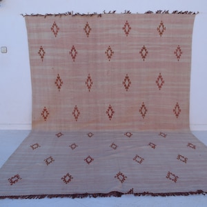 May include: A large, rectangular rug with a light peach and beige color scheme. The rug features a repeating pattern of diamond shapes in a darker reddish-brown hue. The rug is made of a woven material and has a fringed edge.