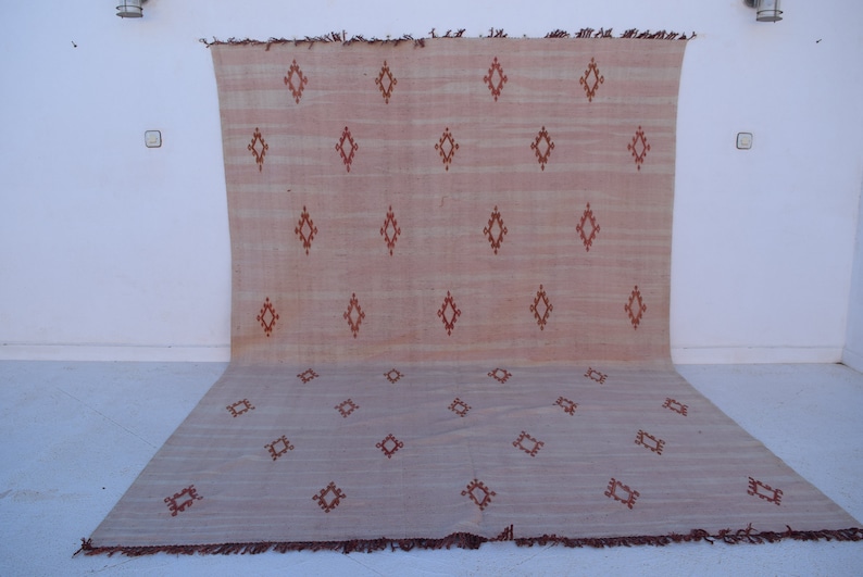 May include: A large, rectangular rug with a light peach and beige color scheme. The rug features a repeating pattern of diamond shapes in a darker reddish-brown hue. The rug is made of a woven material and has a fringed edge.