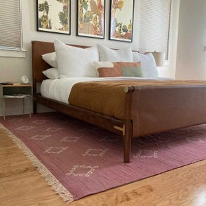 May include: A bedroom scene featuring a bed with a brown leather headboard and frame. The bed is made with white linens and a brown blanket. A pink rug with a geometric pattern is on the floor. Artwork with botanical prints hangs on the wall.