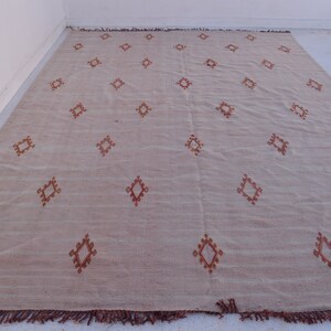 May include: A large, rectangular beige rug with a repeating pattern of diamond shapes in a reddish-brown color. The rug has a fringe along the bottom edge. The rug is made of a woven material.