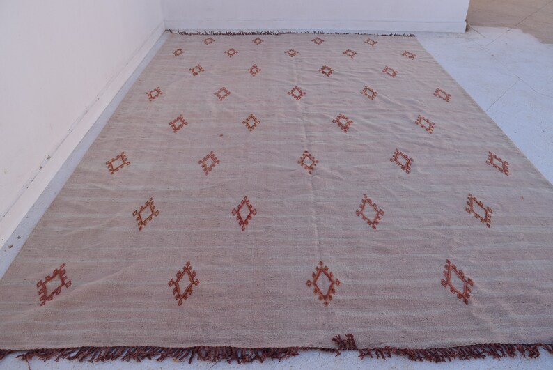 May include: A large, rectangular beige rug with a repeating pattern of diamond shapes in a reddish-brown color. The rug has a fringe along the bottom edge. The rug is made of a woven material.