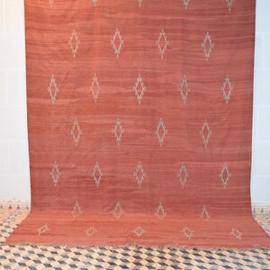 May include: A large, rectangular rug in a warm, reddish-orange hue. The rug features a repeating pattern of diamond shapes in a lighter, neutral color. The rug is displayed against a white wall.