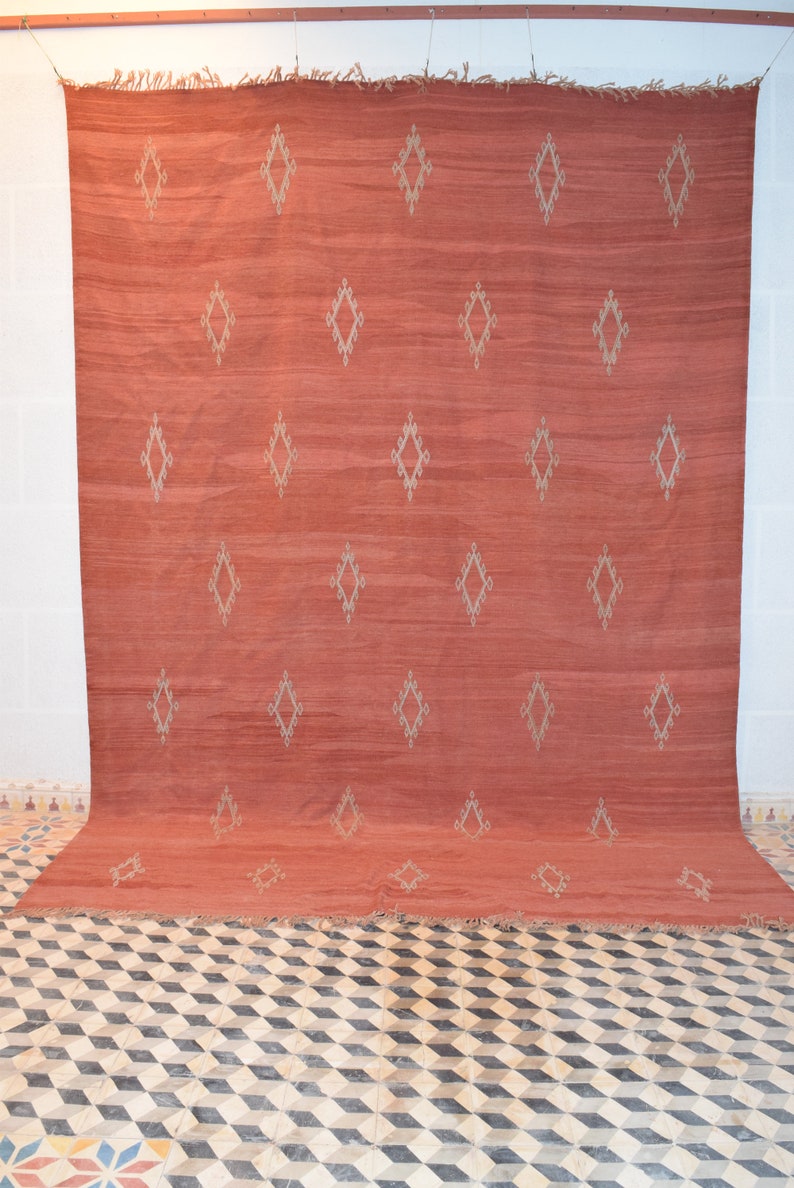 May include: A large, rectangular rug in a warm, reddish-orange hue. The rug features a repeating pattern of diamond shapes in a lighter, neutral color. The rug is displayed against a white wall.
