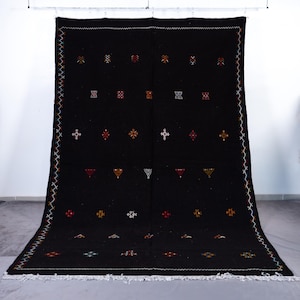 May include: A large, black woven rug with a geometric pattern. The rug features small, colorful embroidered designs in red, yellow, white, and green. The border has a zig-zag stitch in multiple colors, and the bottom edge has a white fringe.