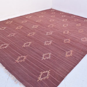 May include: A large, rectangular rug in a deep burgundy hue, featuring a repeating pattern of diamond-shaped designs in a lighter beige color. The rug has a textured appearance with visible vertical lines and fringed edges.