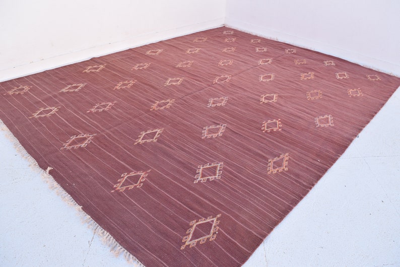 May include: A large, rectangular rug in a deep burgundy hue, featuring a repeating pattern of diamond-shaped designs in a lighter beige color. The rug has a textured appearance with visible vertical lines and fringed edges.
