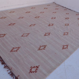 May include: A large, rectangular rug with a light beige background and a repeating pattern of rust-colored diamond shapes. The rug has a fringed edge and is made of a woven material.