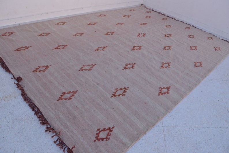 May include: A large, rectangular rug with a light beige background and a repeating pattern of rust-colored diamond shapes. The rug has a fringed edge and is made of a woven material.