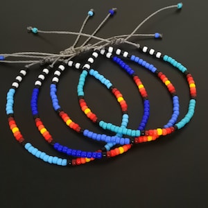 May include: Five colorful beaded bracelets with a black and white beaded bracelet on top. The bracelets are made with small, round beads in shades of blue, red, yellow, and turquoise.
