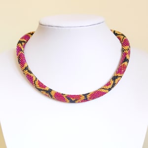 Könnte beinhalten: Eine rosafarbene, schwarze und goldene Perlenkette. Die Kette besteht aus kleinen Perlen, die in einem Muster miteinander verwoben sind.