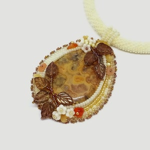 May include: A beaded necklace with a pendant featuring a brown agate stone surrounded by white, brown, and orange beads. The pendant is decorated with small white flowers and brown leaves.