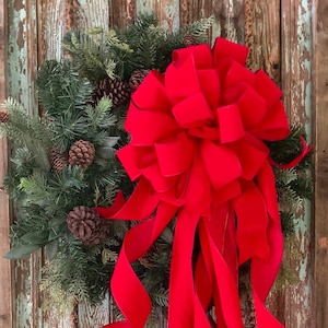 May include: A festive Christmas wreath with evergreen branches, pinecones, and a large, vibrant red velvet bow. The bow's long, flowing ribbons cascade down, adding a touch of elegance to the holiday decor. The wreath is displayed on a rustic, weathered wooden door.