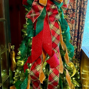 May include: A large Christmas tree topper bow featuring layered ribbons in red, emerald green, and gold. The design includes glittery red fabric, solid green glitter ribbon, and a red, green, and white plaid pattern with long, cascading tails.