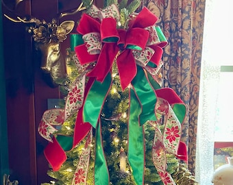May include: A Christmas tree decorated with green and red ribbon, white ribbon with red poinsettia and berry designs, and white lights. The tree is topped with a large red velvet bow with green ribbon.