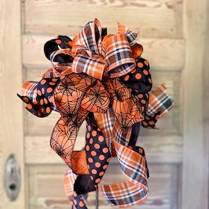 May include: A Halloween-themed bow made with orange and black ribbon. The bow features a spiderweb pattern, polka dots, and plaid. The bow is attached to a metal stake.