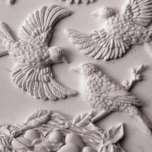 Pode incluir: Escultura em relevo detalhada com v&aacute;rias aves em voo e empoleiradas, juntamente com um ninho contendo ovos. As aves s&atilde;o retratadas com penas e bicos texturizados, e o ninho &eacute; intrincadamente tecido com folhas e galhos. A obra de arte &eacute; monocrom&aacute;tica, esbranqui&ccedil;ada.