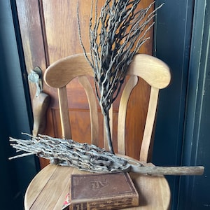 May include: Two bundles of dried brown branches, resembling a witch's broom, are placed on a wooden chair. A small brown book with the title "Deal Stories" is on the chair.