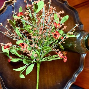 May include: A bouquet of artificial flowers with red and orange blooms, green leaves, and small brown buds. The bouquet is placed on a dark brown, ornate wooden tray. A green glass bottle is partially visible.