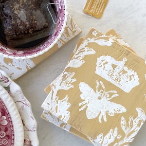 May include: Set of four yellow square coasters with a white bee, crown, and floral design. The coasters are stacked on top of each other.