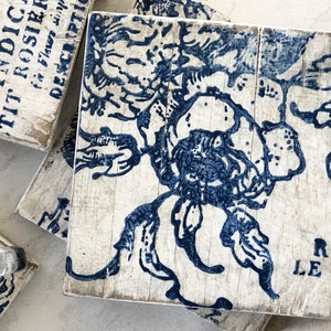 May include: Set of four blue and white floral coasters with a distressed wood finish. The coasters feature a floral design with the words "LE ROI" printed on one of the coasters.