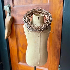 May include: A decorative, natural-looking wreath made of intertwined brown twigs sits atop a beige dress form. The dress form is on a metal stand. The background features a wooden door and a dark wall.