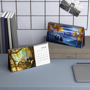 May include: Two spiral-bound desk calendars. One calendar shows the month of August with a colorful illustration. The other calendar displays the text "The Ring Sets Out" with a landscape illustration. Both calendars are on a desk.
