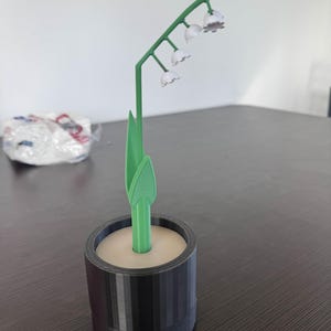Puede incluir: Una planta de lirios del valle impresa en 3D en una maceta negra. La planta tiene un tallo y hojas verdes, con pequeñas flores blancas en forma de campana. La maceta es cilíndrica con líneas verticales.