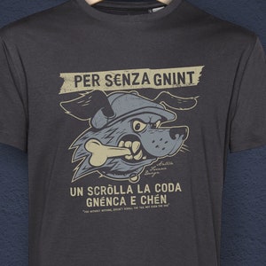 Könnte beinhalten: Dunkelgraues T-Shirt mit einem Cartoon-Hund-Motiv. Der Hund, der einen Hut trägt und einen Knochen hält, ist das zentrale Designelement. Italienischer Text lautet "PER SENZA GNINT" und "UN SCROLLA LA CODA GNÉNCA E CHÉN". Das Shirt hängt an einem Holzbügel.