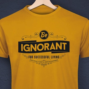 Könnte beinhalten: Senfgelbes T-Shirt mit dem schwarzen Text "Be IGNORANT for successful living", verziert mit dekorativen Schnörkeln. Das T-Shirt hängt an einem Holzbügel vor einem dunkelblauen Hintergrund.